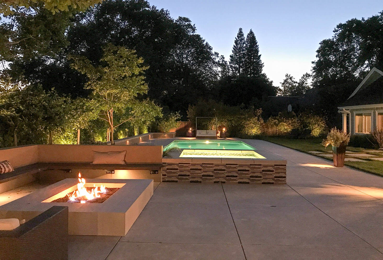 Illuminate - well-lit pool and Jacuzzi with rectangular fire pit, and lights under wrapped seating section, and lights coming from some flower pots as well