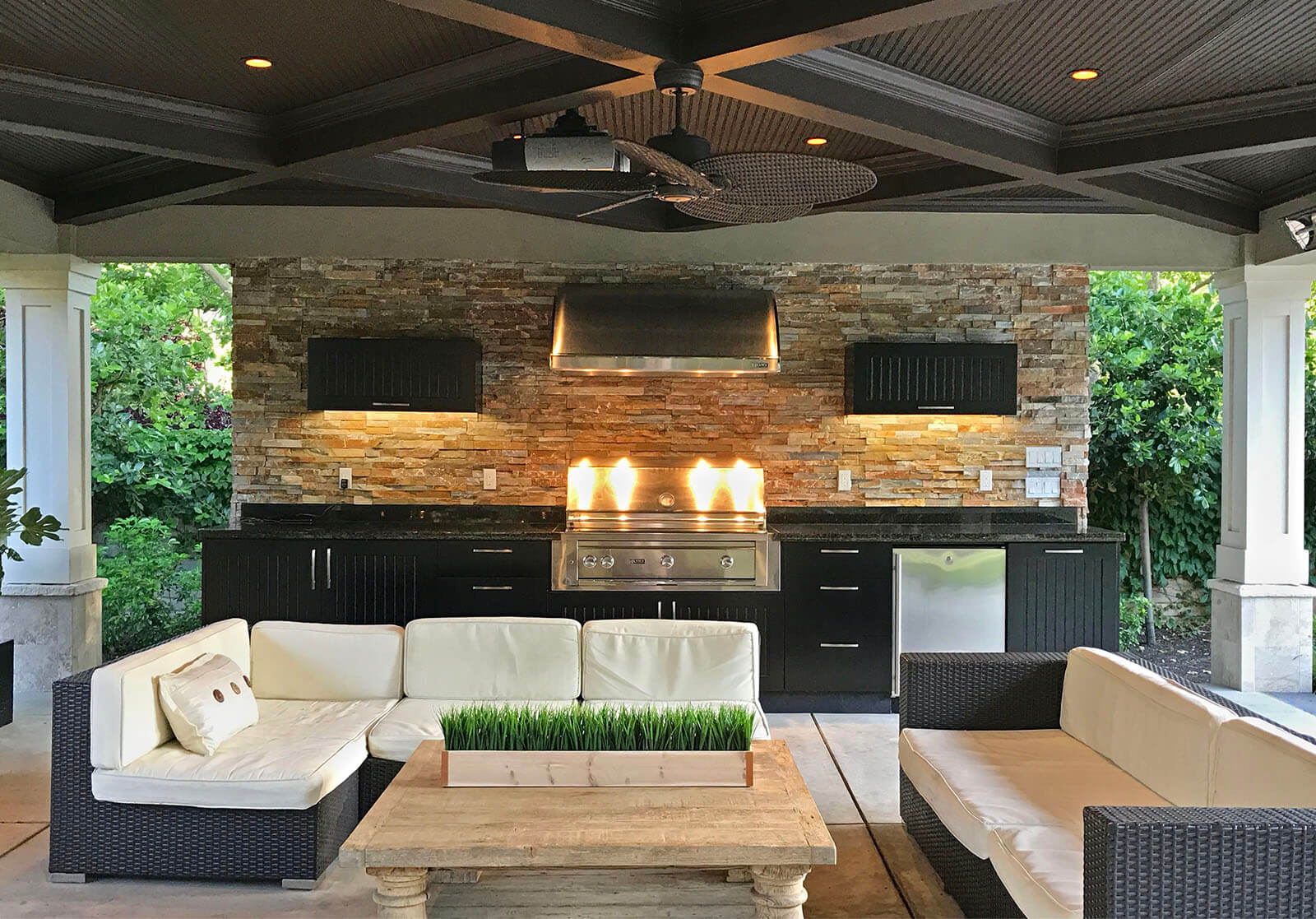 Covered outdoor kitchen and lounge with black granite counters, stone accent wall, stove and hood with contemporary styling