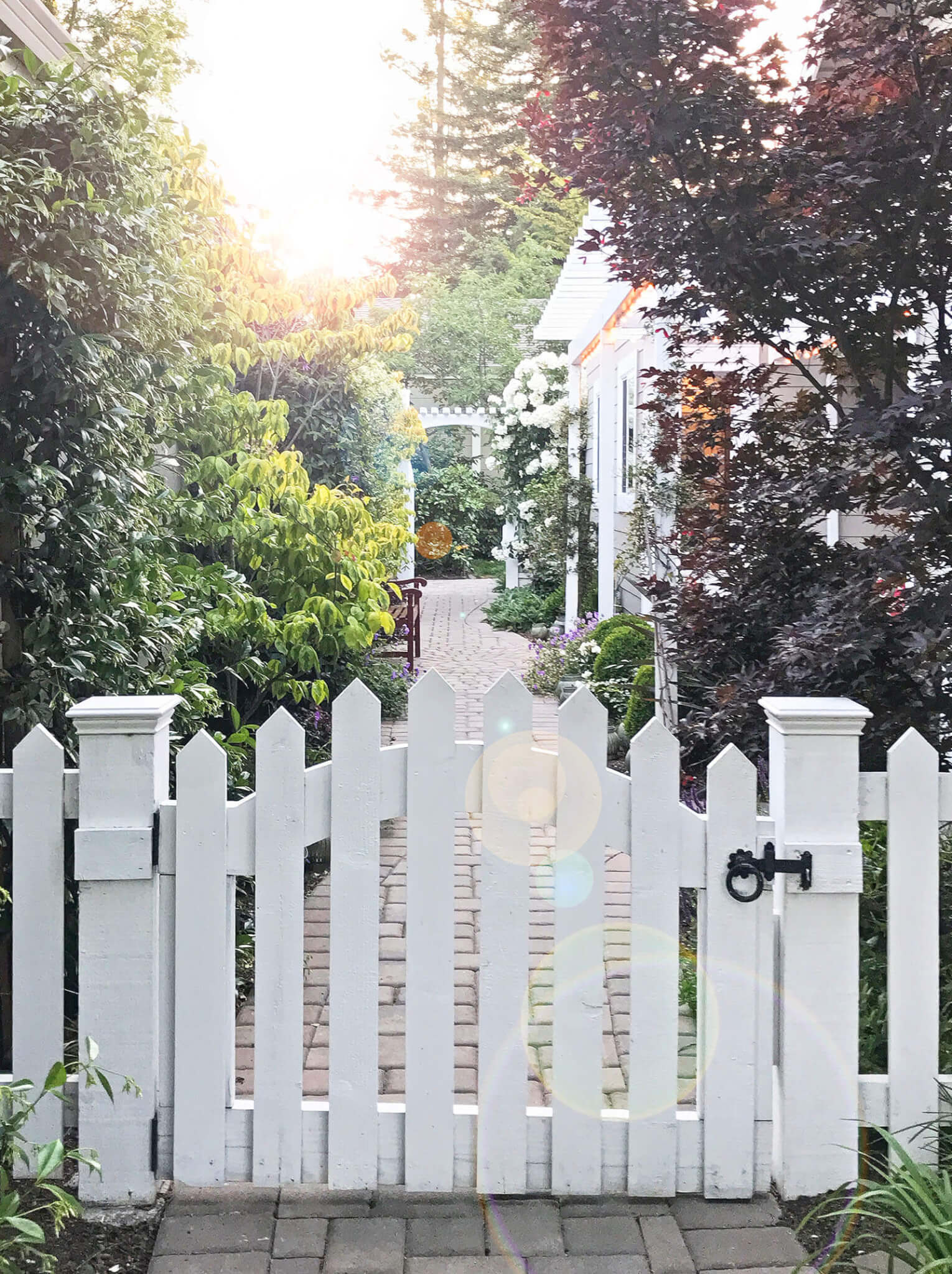 White picket fence gate with wrought iron latch, leading to brick path going to side yard