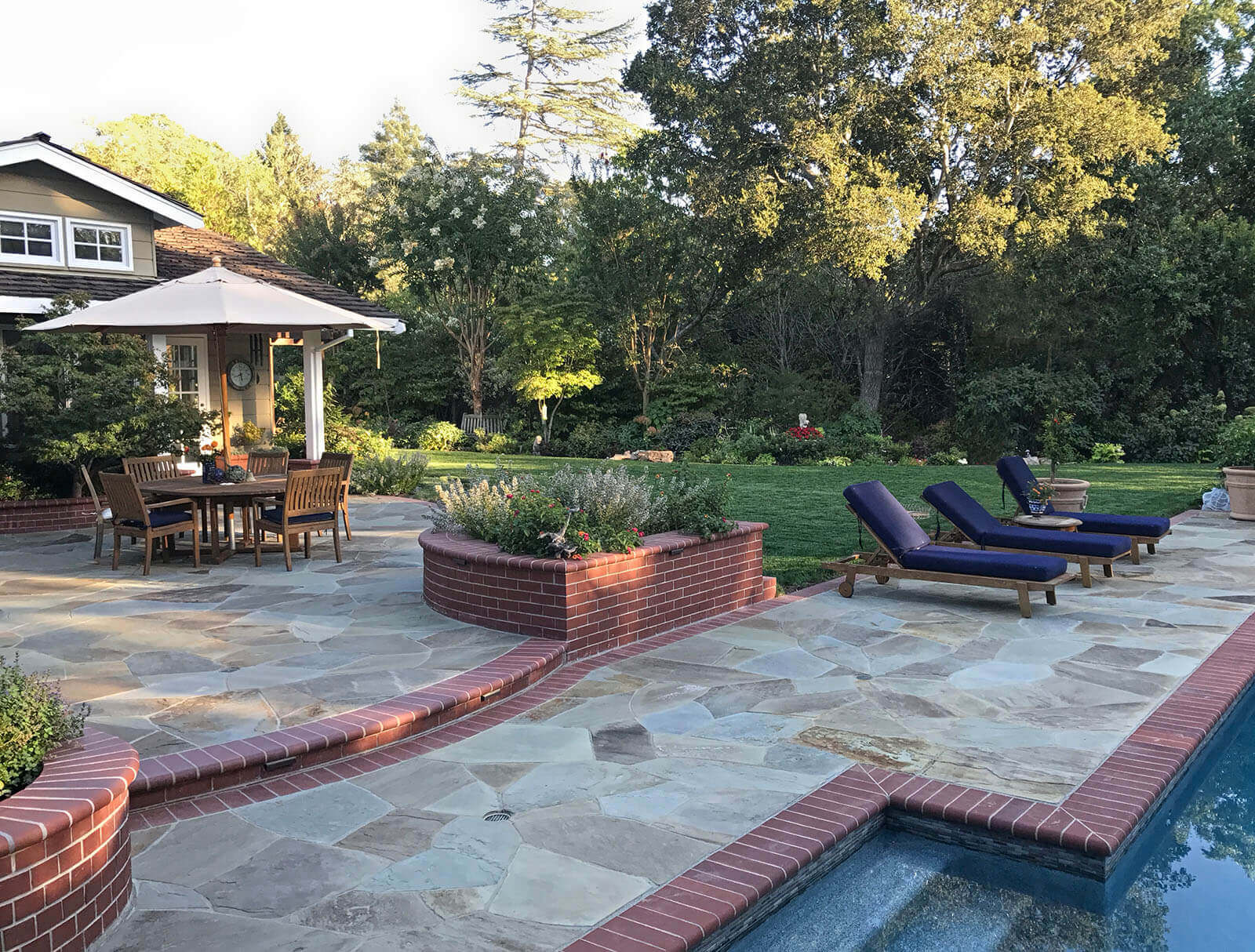 Stone dining patio with brick detailing, poolside lounge, and expansive lawn
