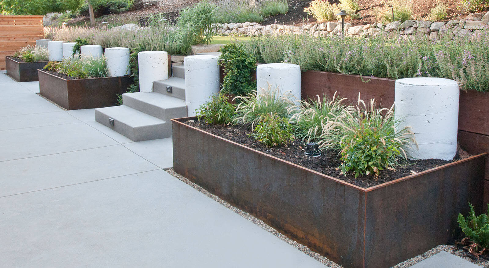 Metal raised beds, with concrete pillars and stairs, leading to elevated lawn and flower beds, with sloped back yard above