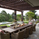 Pergola-covered stone dining patio with rustic table and rattan chairs and chandeliers