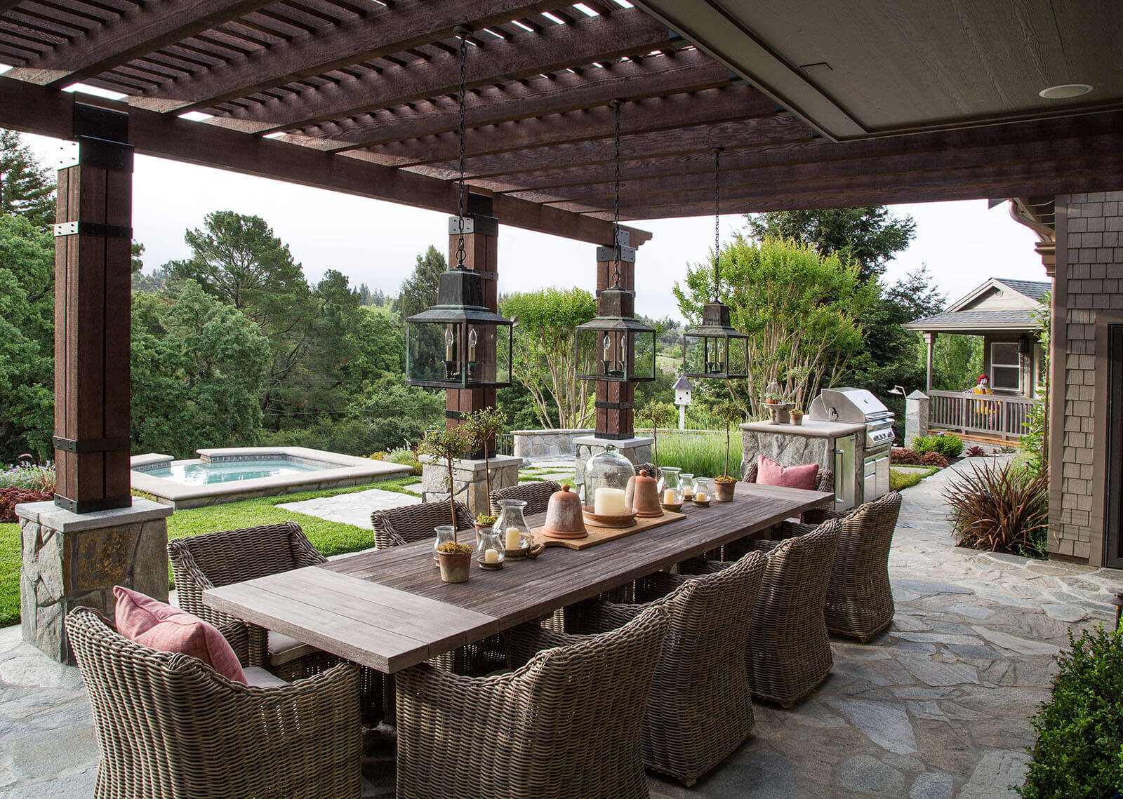 Pergola-covered stone dining patio with rustic table and rattan chairs and chandeliers
