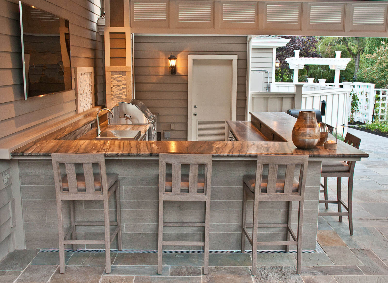 Covered outdoor kitchen with marble counters, grill, sink, and bar seating in a contemporary farmhouse style