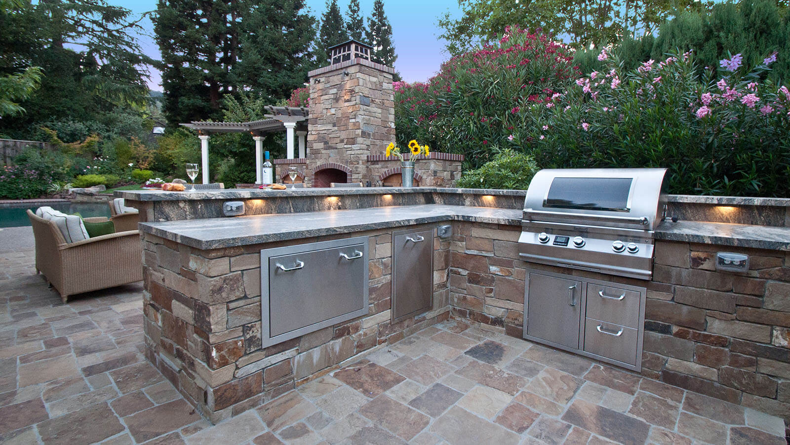 Outdoor kitchen on stone patio with under-counter lighting, built-in contemporary grill, stone fireplace and terraced landscape