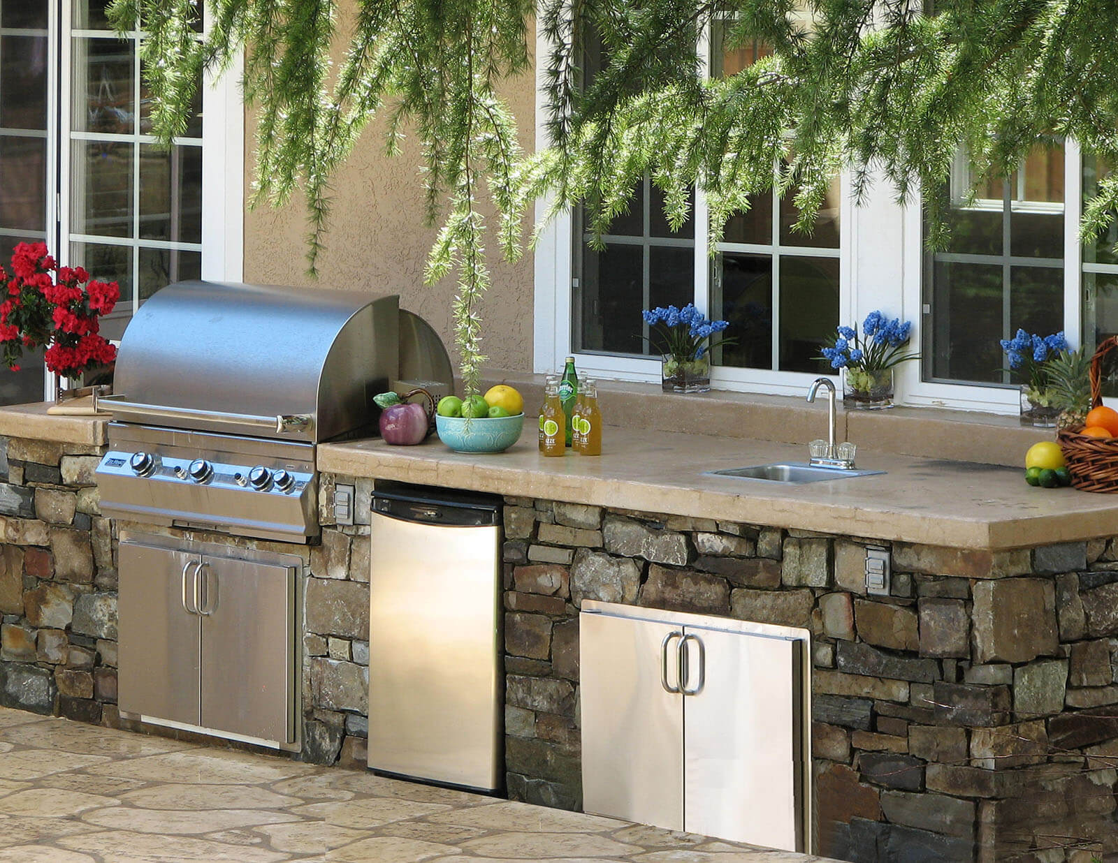 Outdoor shaded grill area with tan stone counter tops, stow space, sink, and stone tile siding.