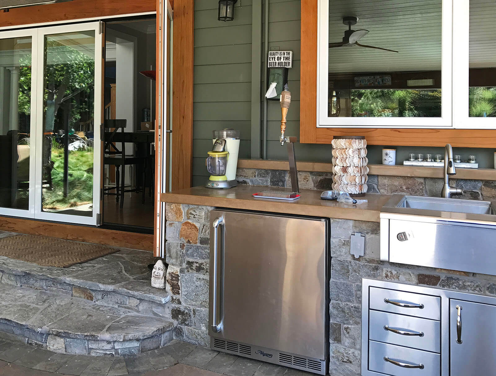 Outdoor kitchen with stainless steel fridge, beer tap, sink and storage, concrete counters and stone detailing