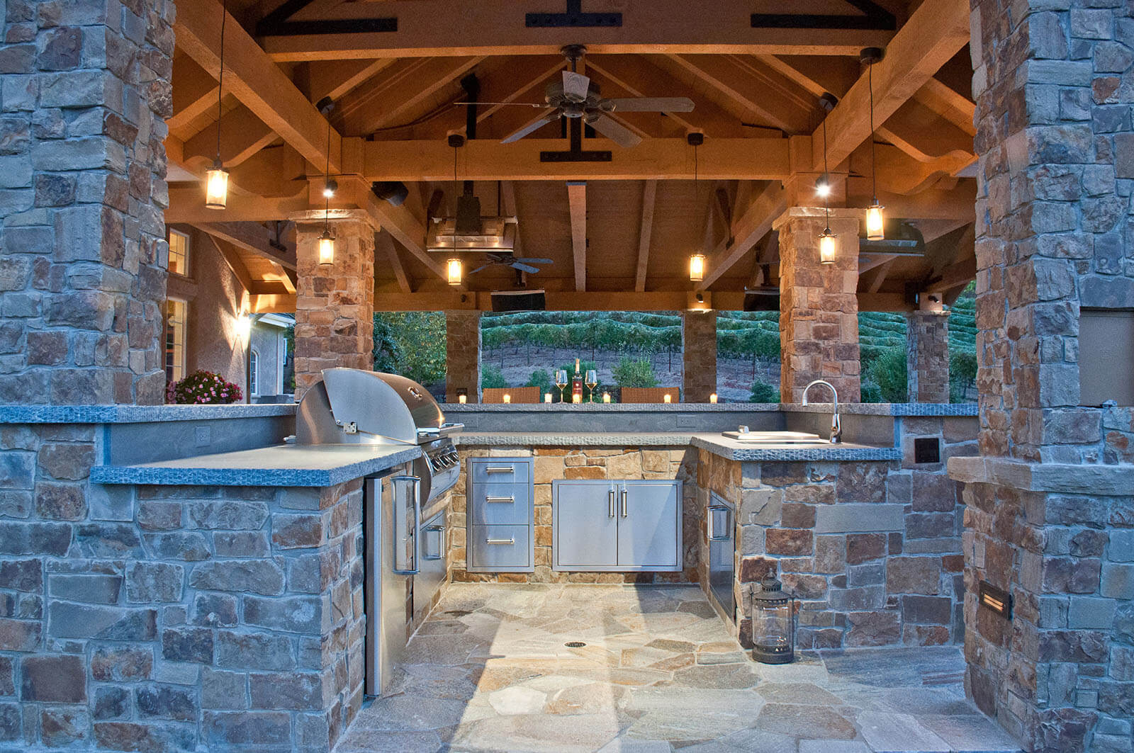 Sonoma-style outdoor bar and grill under wood pavilion with stone accents and romantic lighting, vineyard beyond