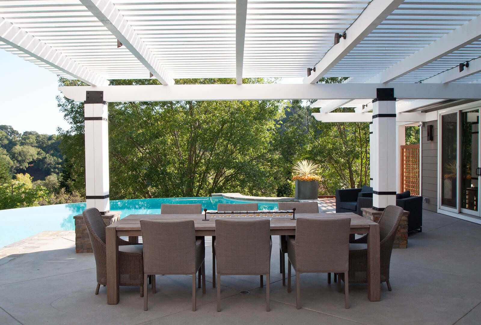 Outdoor dining area under a white pergola with black iron details overlooks curved infinity pool