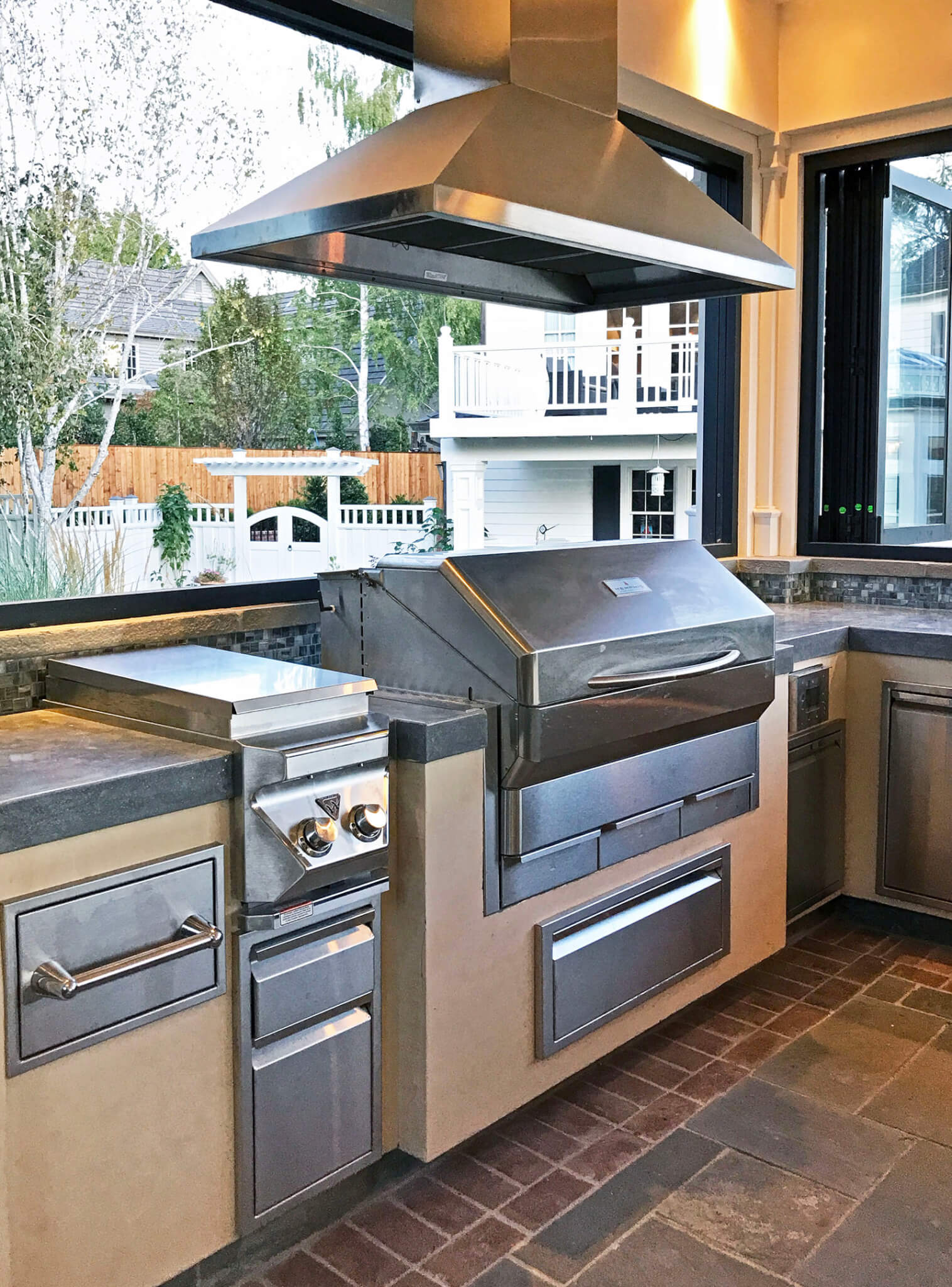 Protected indoor-outdoor kitchen with concrete countertops, built-in contemporary grill with hood, full kitchen amenities and retracting windows