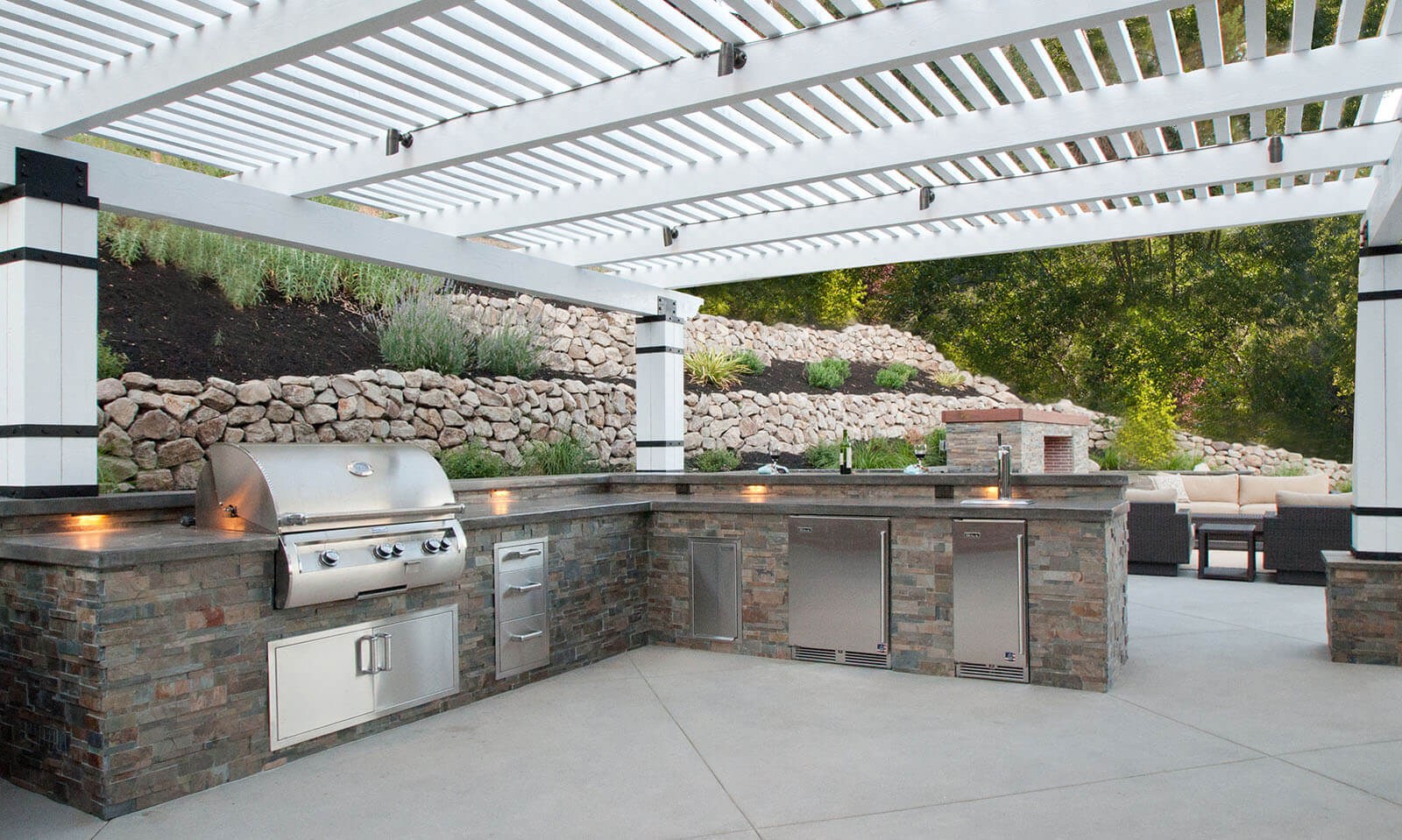 White pergola slat roofed patio with large stone counter-tops and kitchen area with storage, custom grill, and temperature controlled storage areas