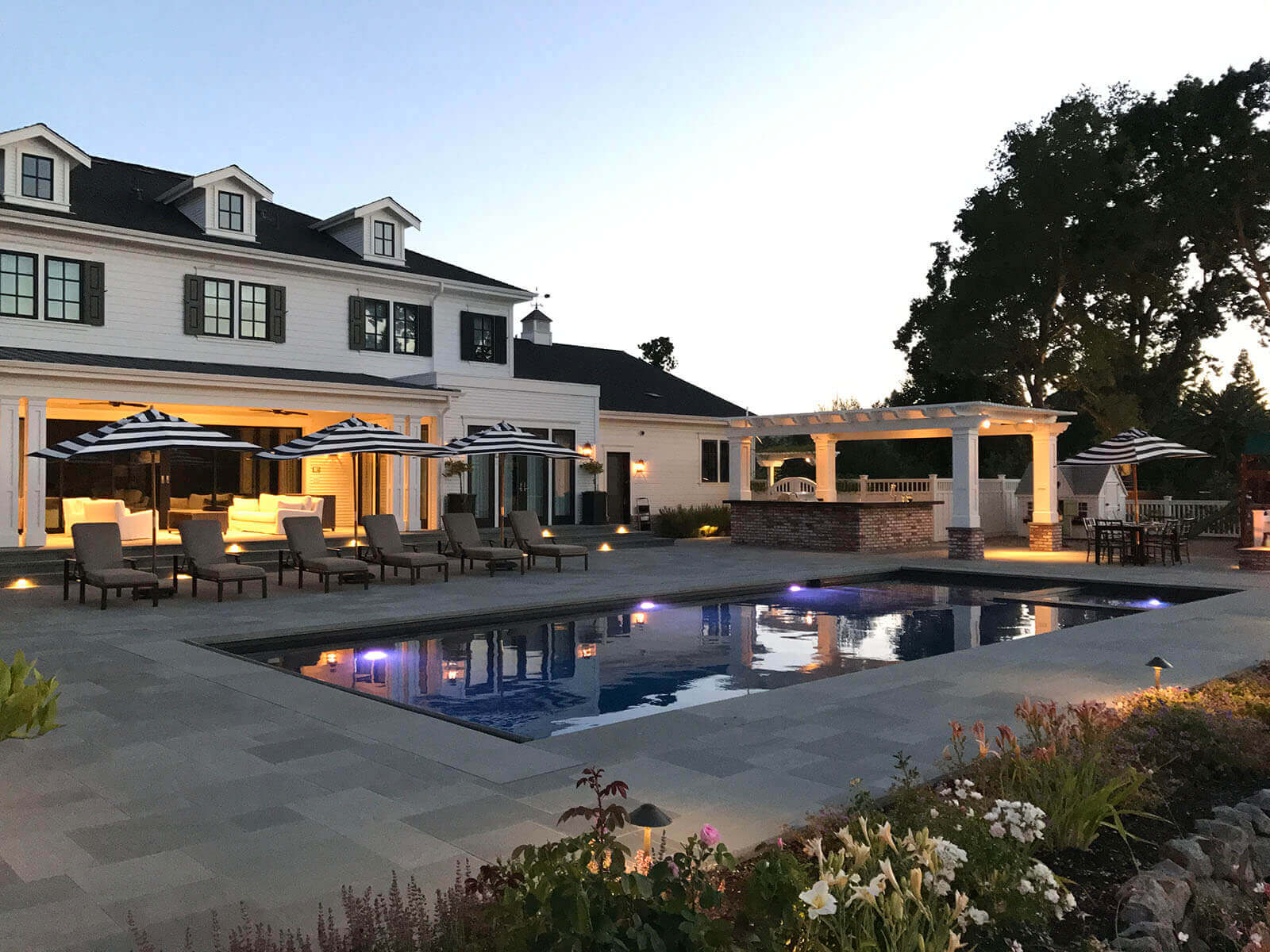 (pool view) Outdoor lighting at sunset, light fixtures around the edge of the structures, and accent lighting on staged gardening areas
