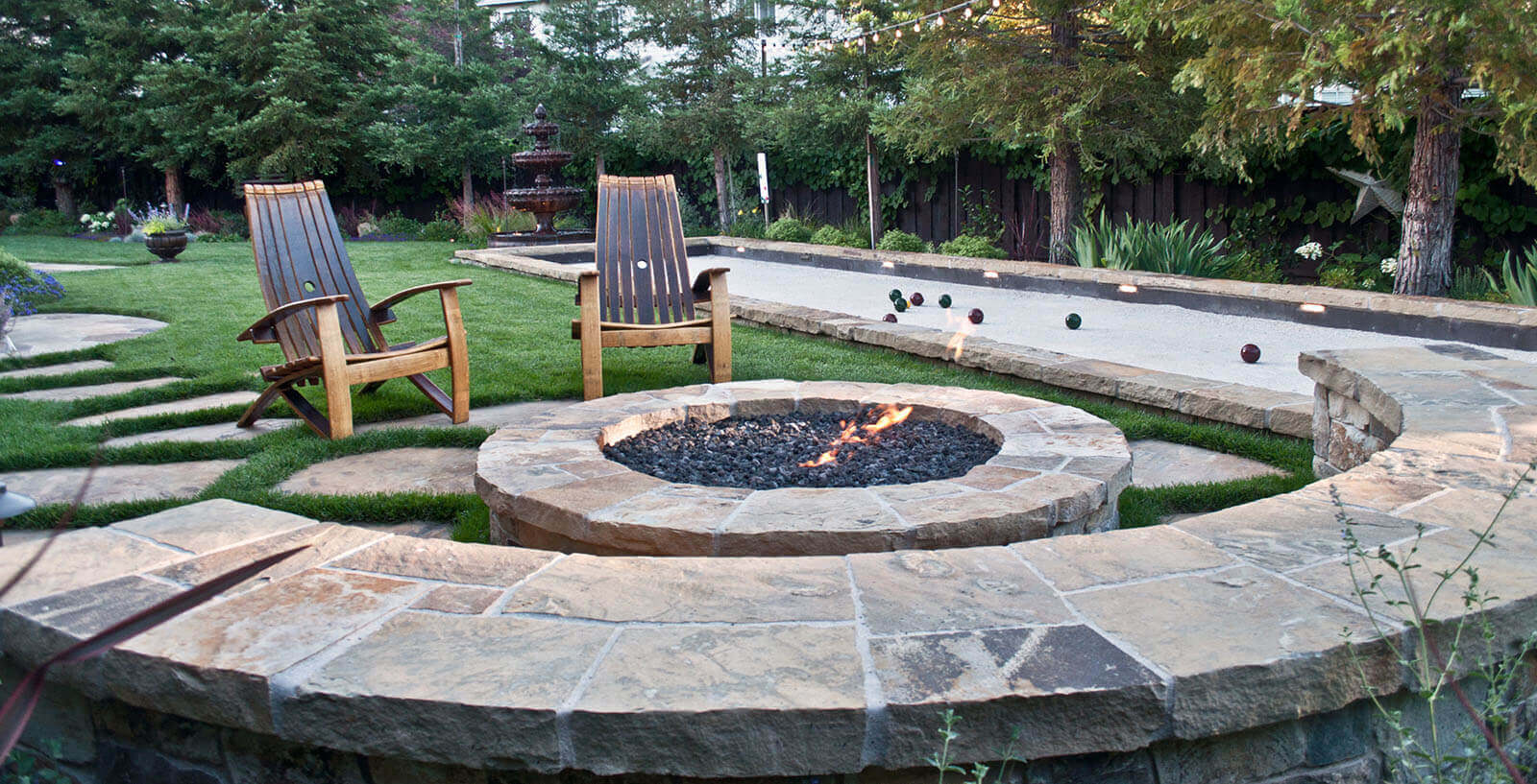 Rounded stone-lined river rock fireplace with rustic wooden lawn chairs and stone wrapped seating overlooking stone-lined lit bachi ball court