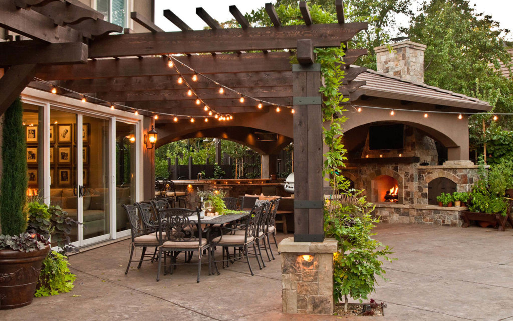 Clay colored tiled patio with tan stone and brick hearth fireplace with veranda covered bbq cooking area with outdoor wrought iron dining set under rustic wood pergola and warm stringed bulb lighting