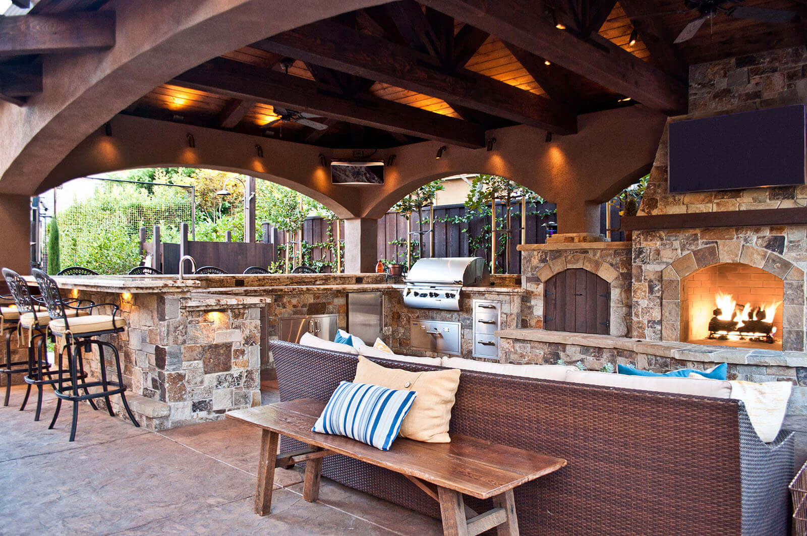 large wrapped stone counter bbq and cooking area connected to a stone mantle fireplace and wood storage area, open bar seating on kitchen counter, and outdoor rattan couch in front of the fireplace. The whole building is covered with accent lit curved archway roofing made from stone and dark wood