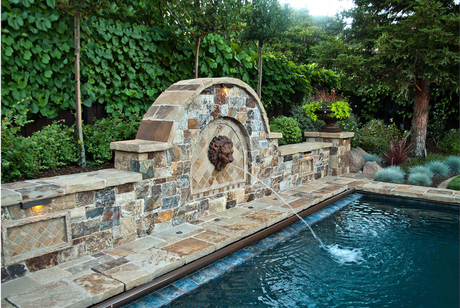 Lavish stone tile wall with curved top centerpiece with lions head fountain leading into the far end of a dark azure, stone tile lined swimming pool