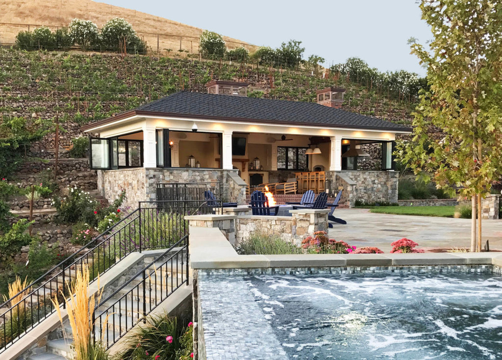 flowing mosaic tile waterfall with wrought iron railing on stone stairway leading away from round stone fire pit surrounded by blue lawn chairs, and behind it, a covered lounge area with open bar and kitchen area