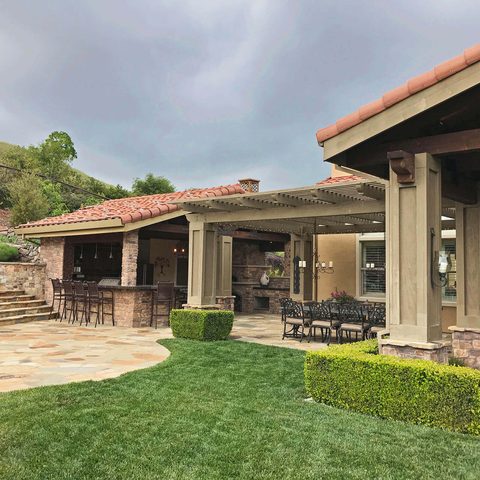 Tiered veranda with slatted roof and connected all-weather structures with Mission-style roof tiles in a landscape with a warm sandstone patio, lawn, and traditional boxwood hedge