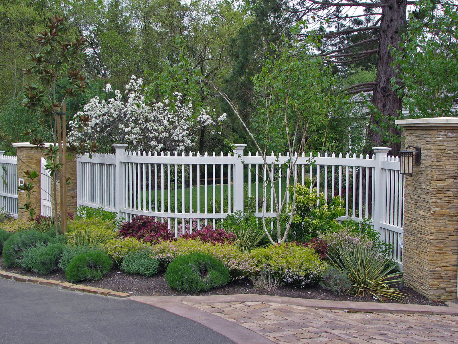 White thin picket fence leading between stone tile columns along side paved driveway with small flowering shrubs and plants
