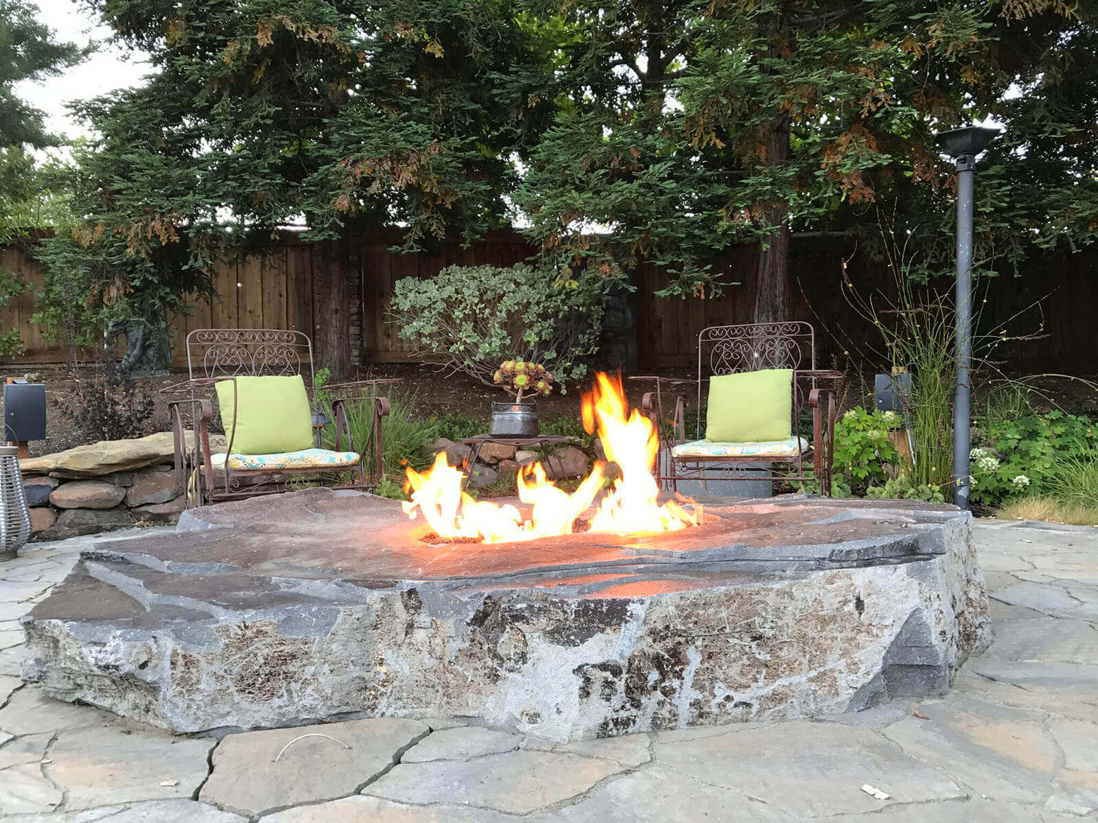Stone slab fire pit in casual outdoor living area with wrought-iron chairs and landscaped garden