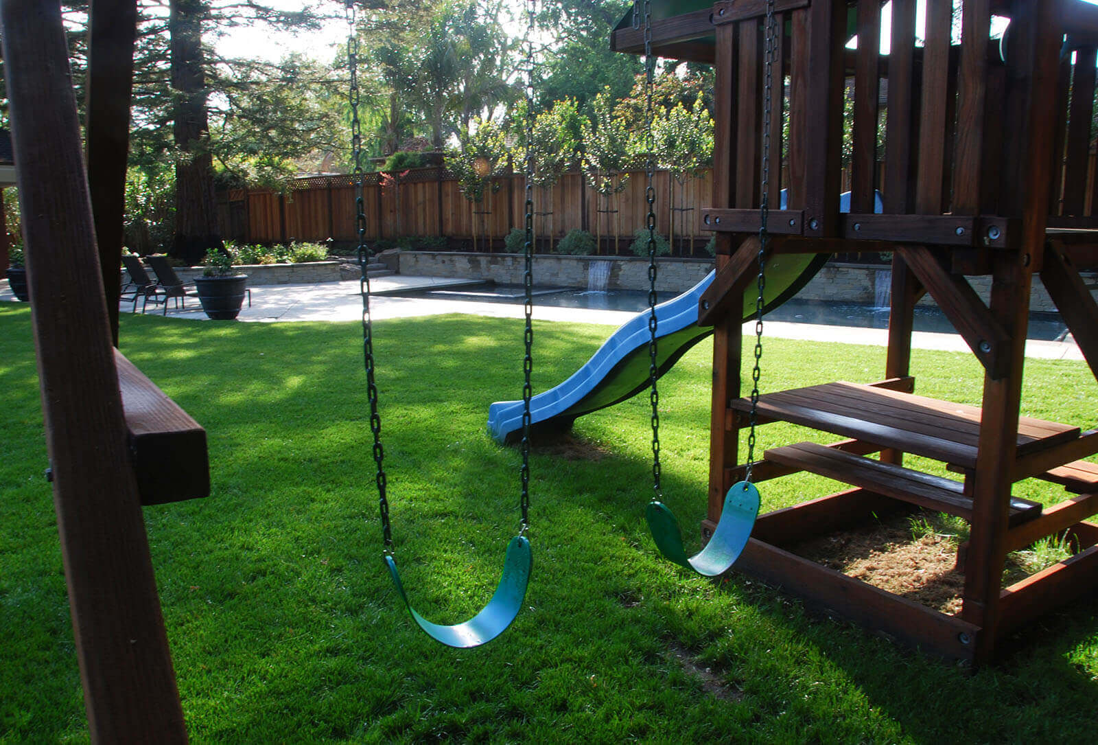 Little jungle gym with wavy slide, climbing fort, and two chain swings in soft lush shaded yard with reflection pool and stone tile patio