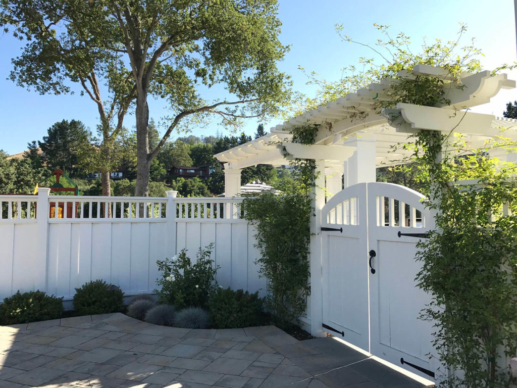 Bright and sunny view of double door gate with wrought iron fixtures and pergola being covered by a climbing rose bush with white fence wrapping around a stone tile patio