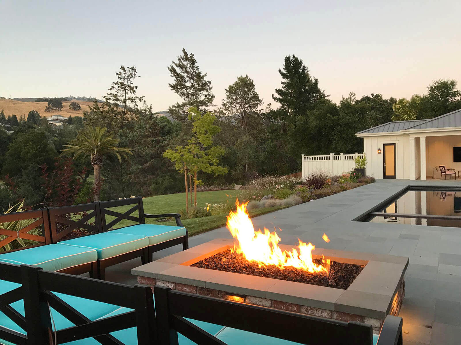 Black outdoor sectional with light blue accent cushions surrounds a low rectangular gas firepit. The bluestone patio and ultra-contemporary pool and spa beyond look out on a California-style landscape.