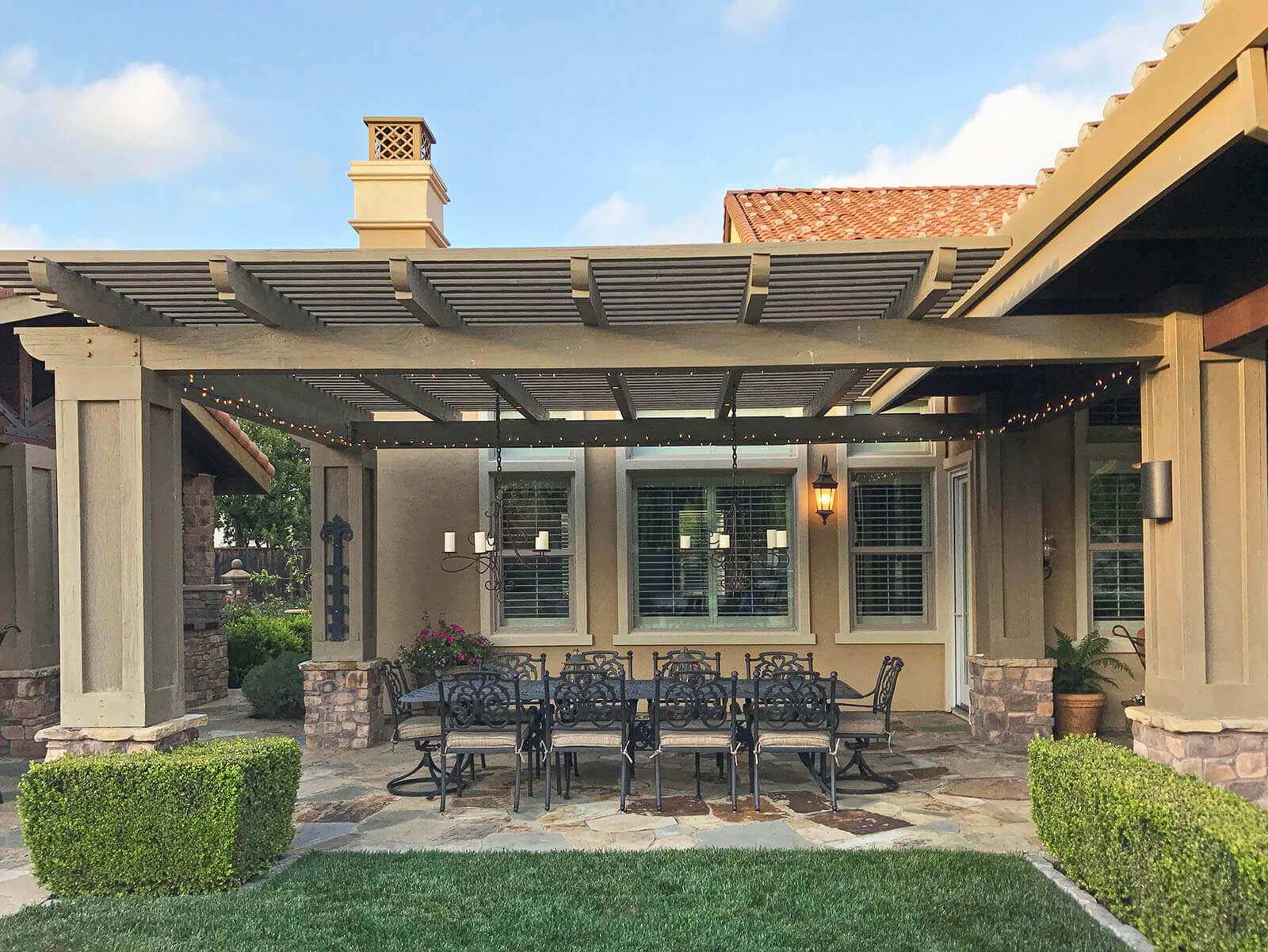 Wood slat-roofed pergola with stone detailing on a stone patio with wrought iron dining furniture and chandeliers