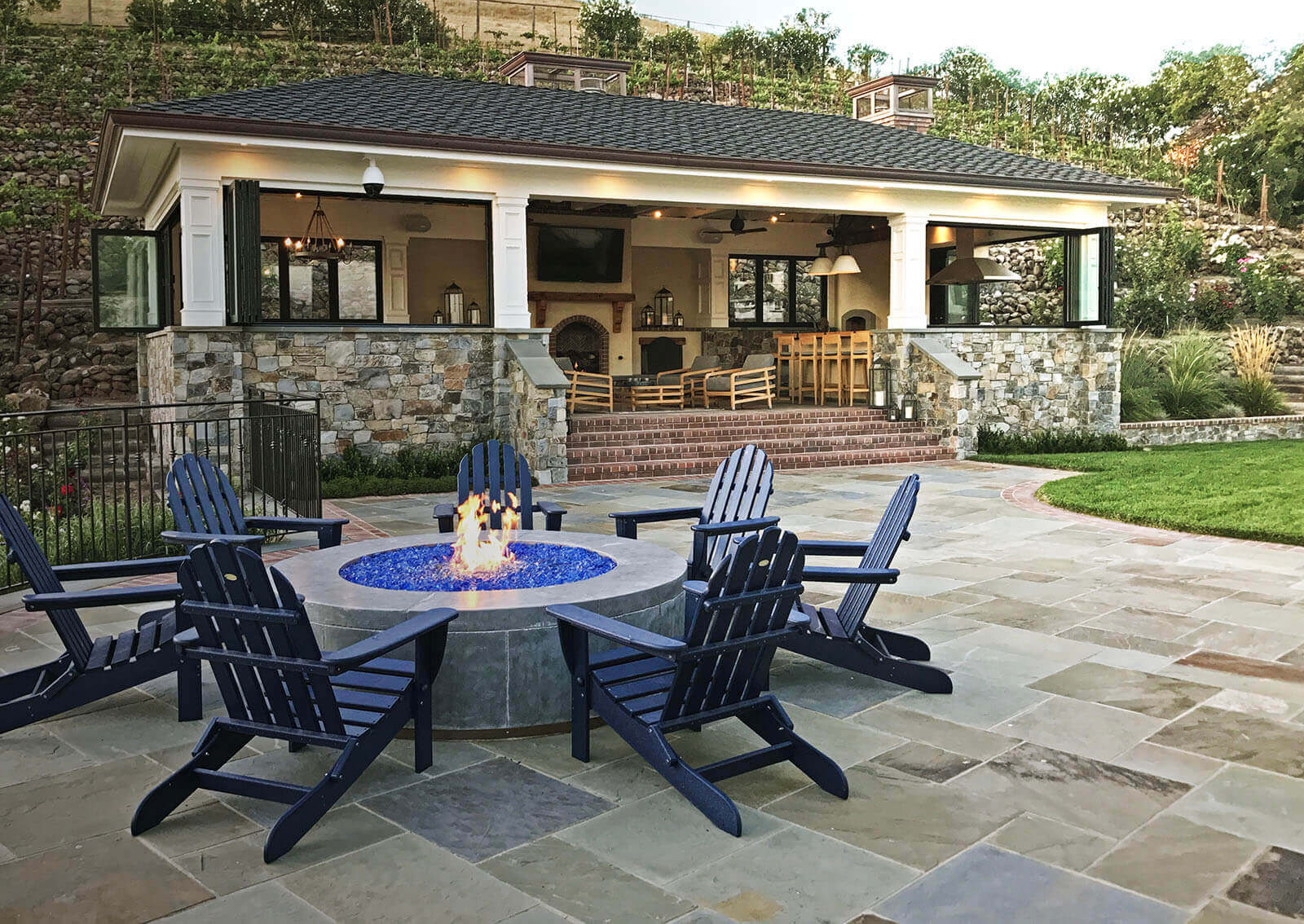 Transitional concrete and stone fire pit with blue crushed glass and Adirondack chairs on stone patio with cabana behind.