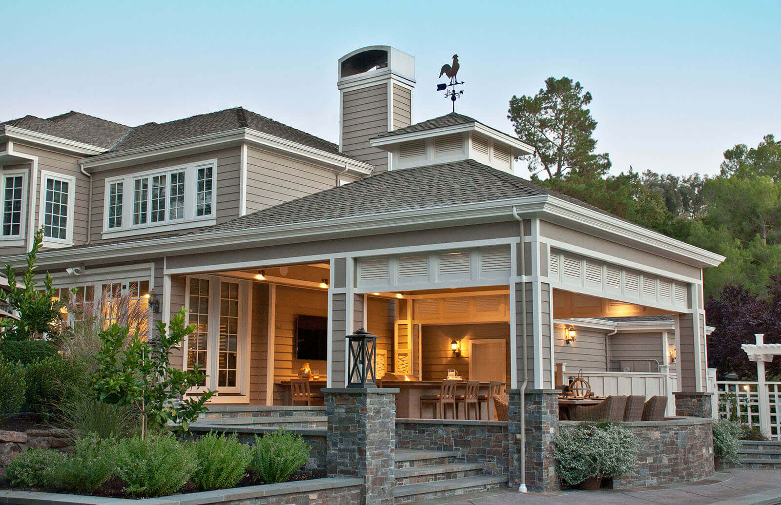 Contemporary farmhouse-style covered veranda with ambient mood lighting, dining area, and kitchen on terraced patio with steps leading down to the landscape