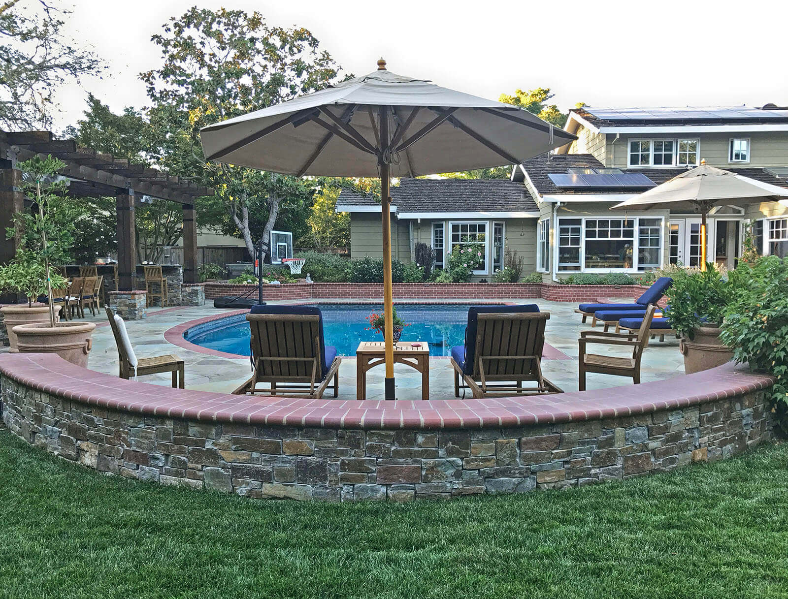 Brick-accented pool with surrounding stone tile patio, poolside pergola and lounge