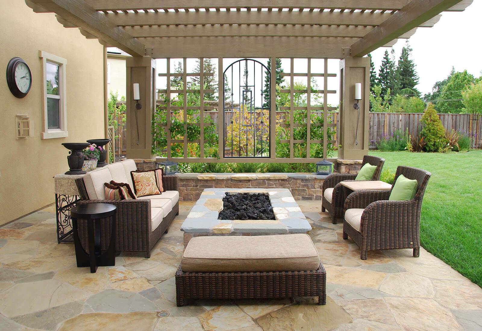 Wood slat-roofed pergola with trellised vines, custom wrought iron panel, and sconce lighting shelters a low stone firepit and lounge seating on a flagstone patio