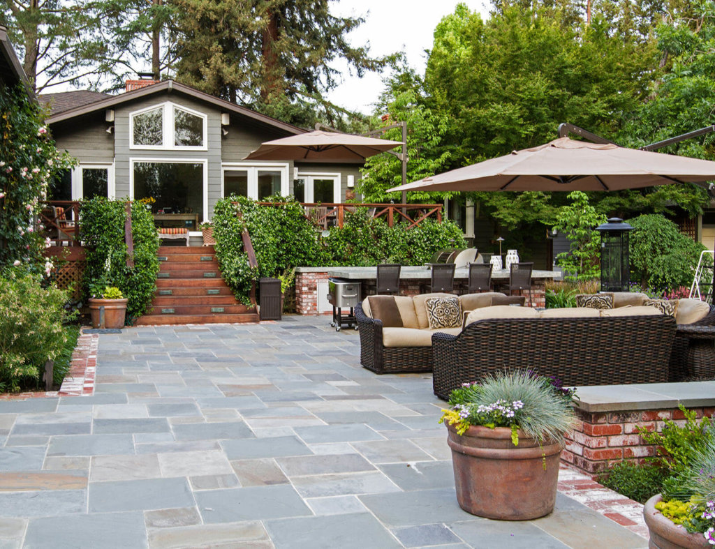 brick-lined slate tile open patio with stone topped bench seating area, rattan couch lounging area (with shaded umbrella) and stone table dining set with wrought iron chairs, further back are dark wood stairs with vine taken railings to an elevated back deck