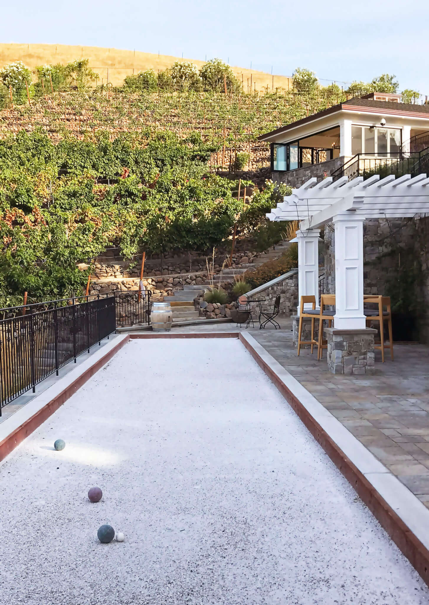 White tile-lined bocce-ball court, next to white pergola, looking up to vineyard on the hill