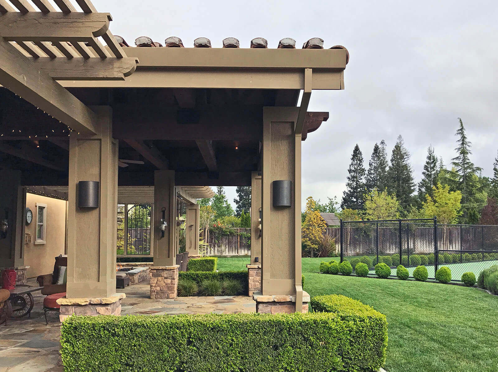 Tiered veranda with slatted roof and connected all-weather structure with Mission-style roof tiles and metal sconce lighting shelters a stone patio with traditional boxwood hedge