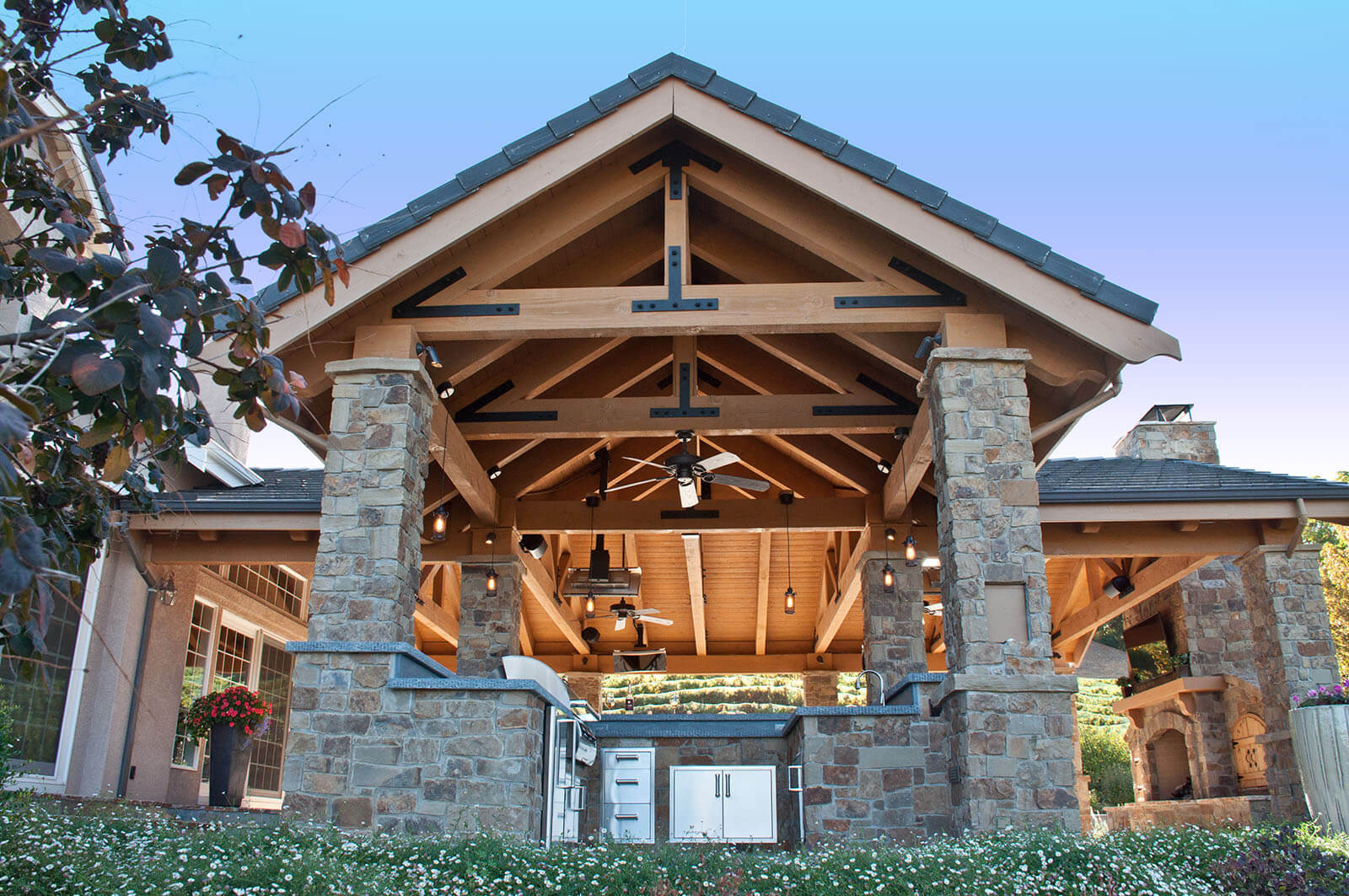 Stone pillars support a complex wooden pergola with open rafters, outdoor kitchen and fireplace