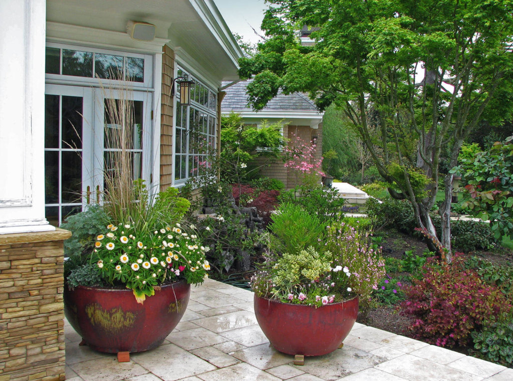 Large ceramic red flower pots with blooming plants on white stone patio overlooking lush landscaped yard with trees and flowering bushes