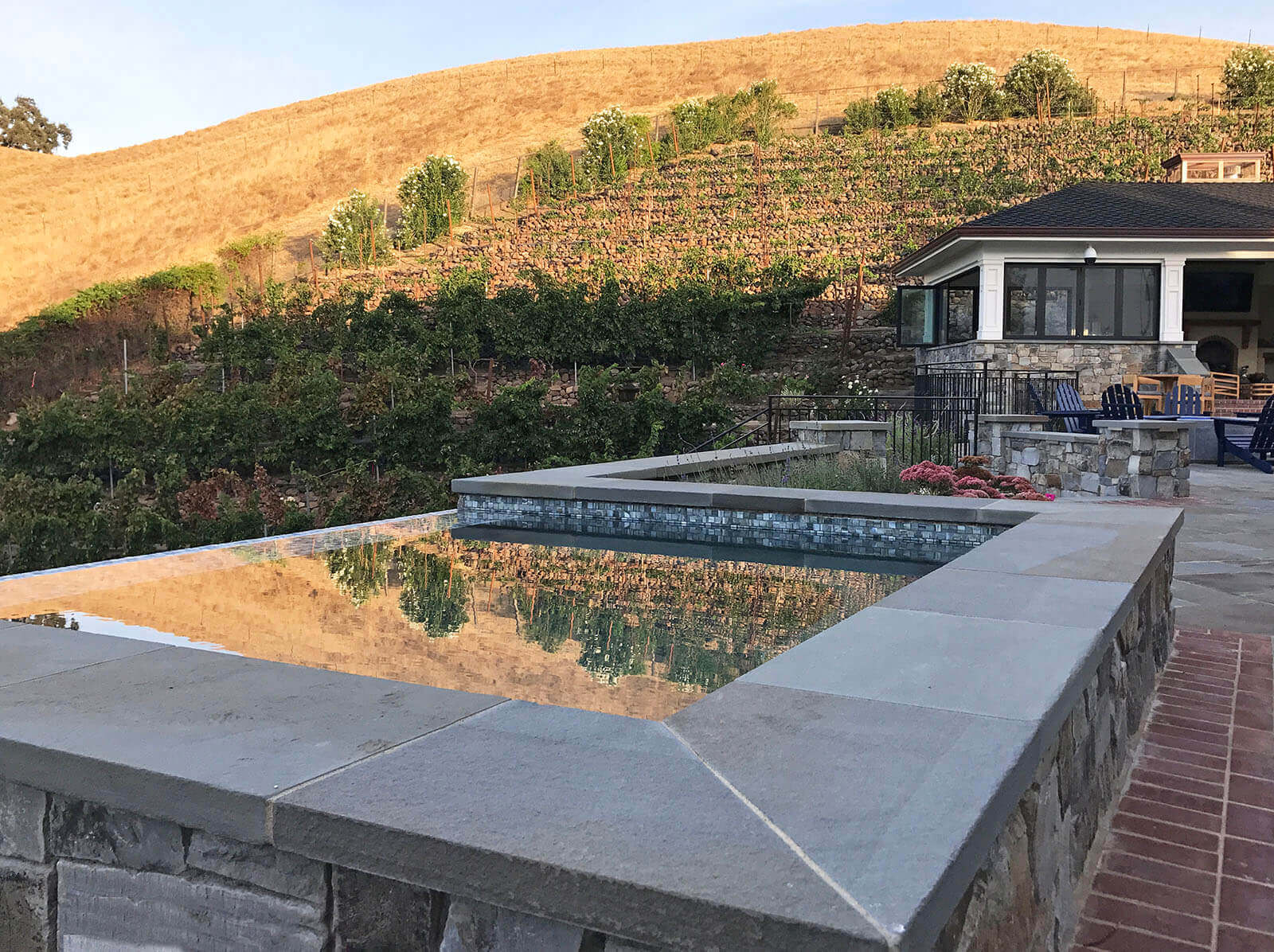 Infinity-edge square spa with bluestone seatwall reflects a hillside vineyard