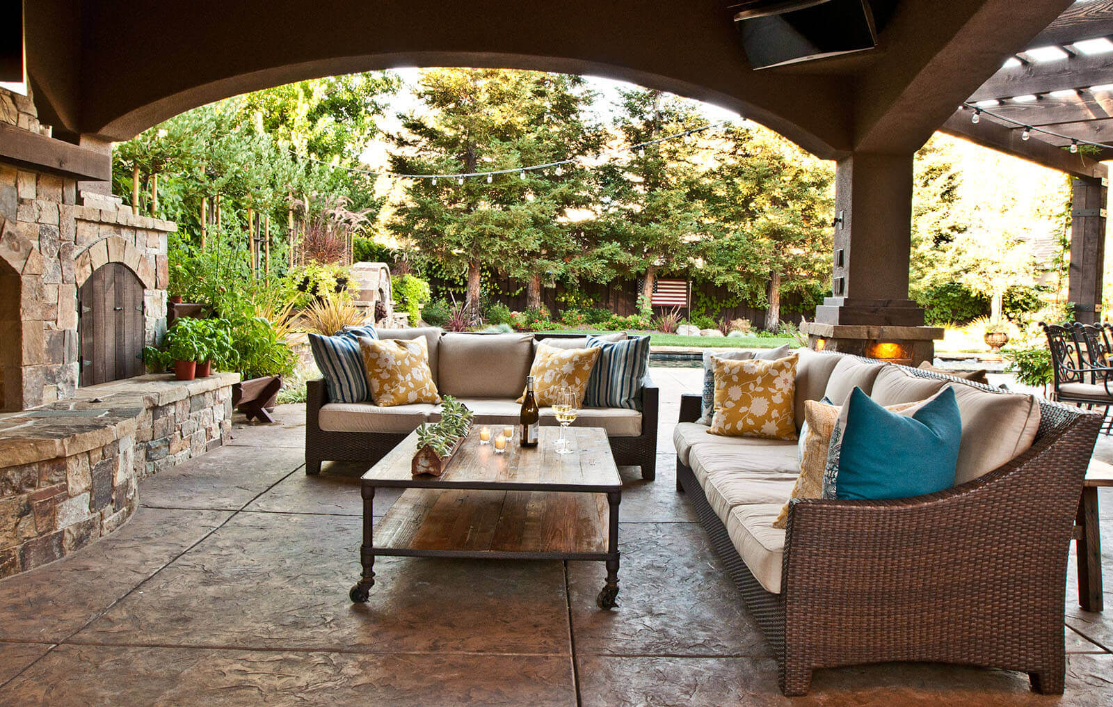 Arched pergola covers an outdoor lounge on rustic tile patio with view of tree-lined landscaped yard
