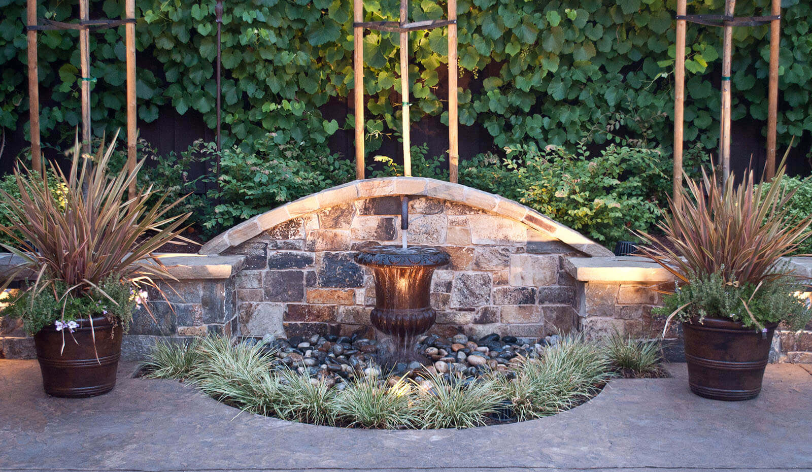 Small potted fountain with stone backdrop and accented pots with grass and plants inside