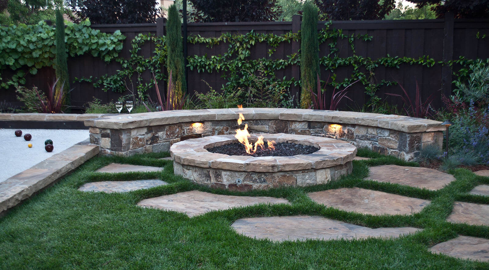 Low round stone fire pit with curved seat wall on casual flagstone patio set in lawn