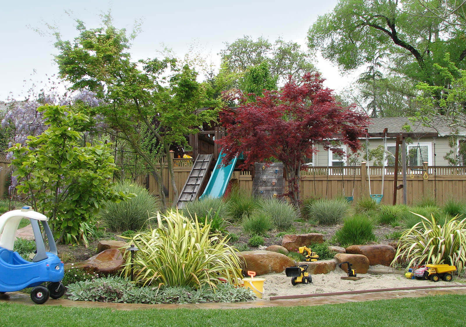 Play - ornamental grass plants spread evenly in yard with stone-lined sandbox, a jungle gym slide with two swings and a swing bar