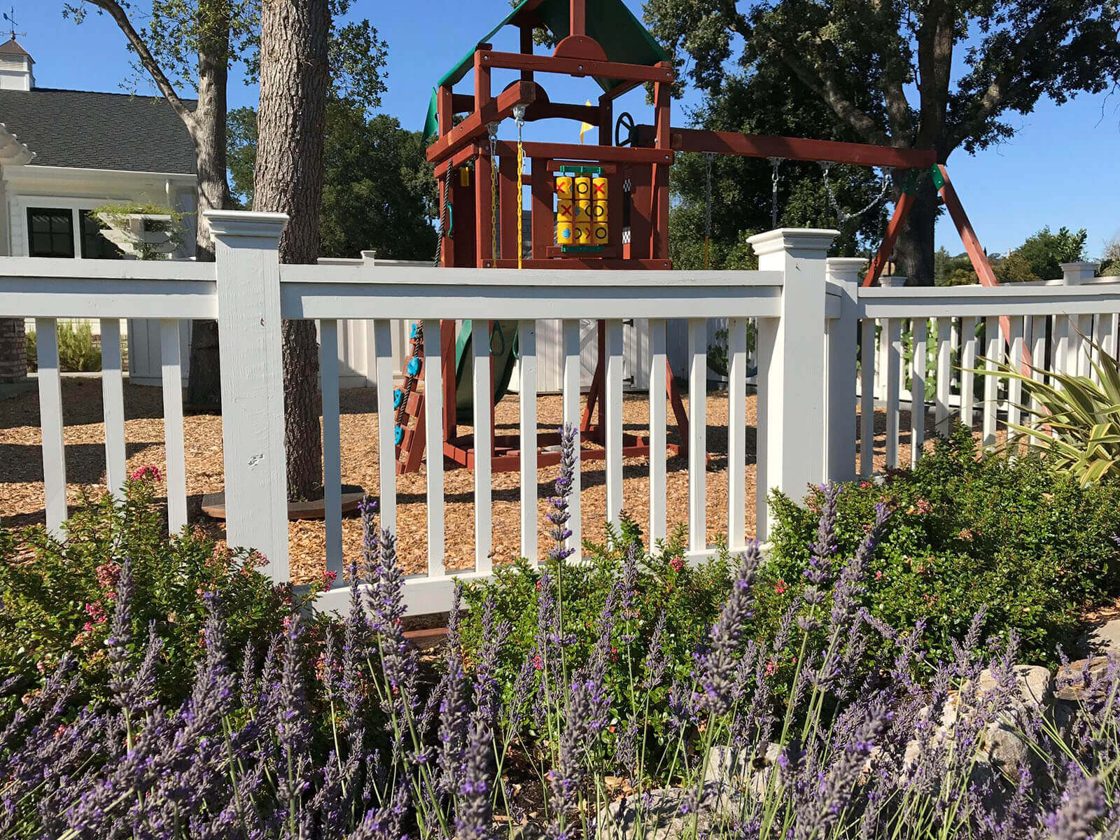 mulch covered play area with tall trees, a full jungle gym (with tic-tac-toe!) and swing set surrounded with white slat fencing and square topped fence posts, with lavender and shrubery outside