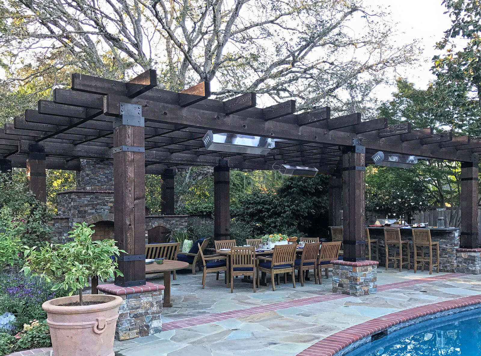 Rustic wood pergola with stone and brick detailing shelters an outdoor dining area with stone fireplace and outdoor bar, on a poolside patio with matching stone and brick details