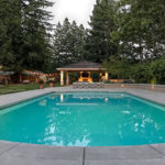 Contemporary pool in concrete patio with low wall and accent lighting, covered pergola and lush lawn