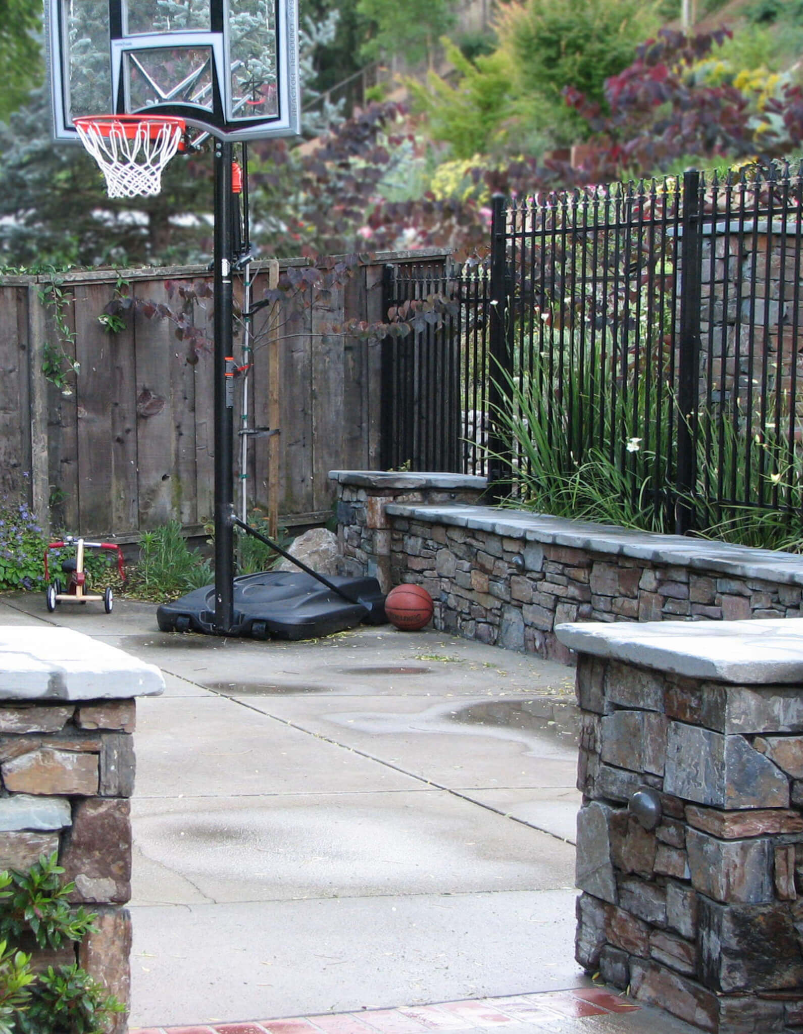 Concrete deck play area with basketball hoop, lined with tile-topped rock walls and rod iron fence.