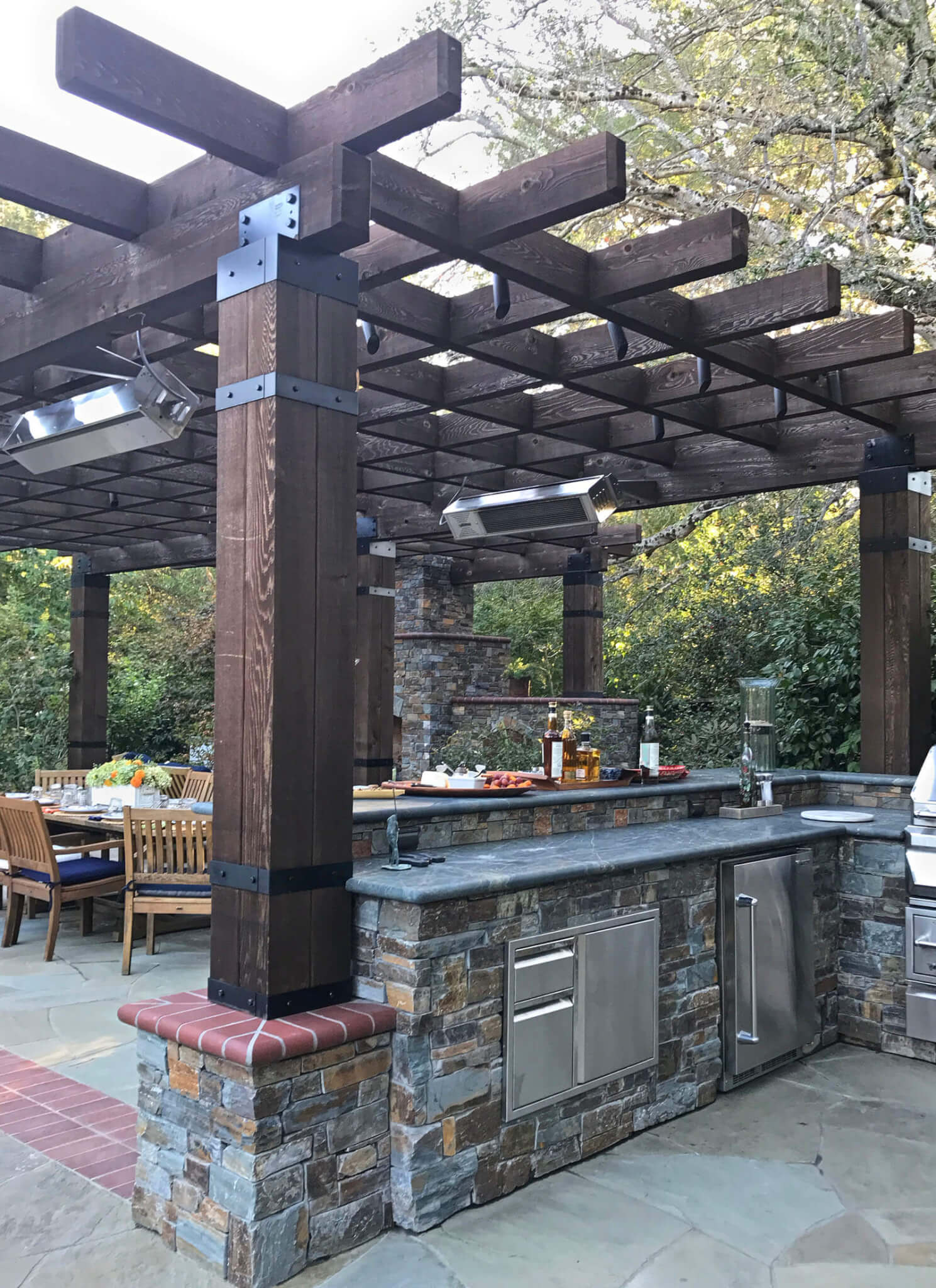 Rustic wood pergola with brick detailing shelters an outdoor dining area with stone fireplace and tiered stone outdoor kitchen and bar.