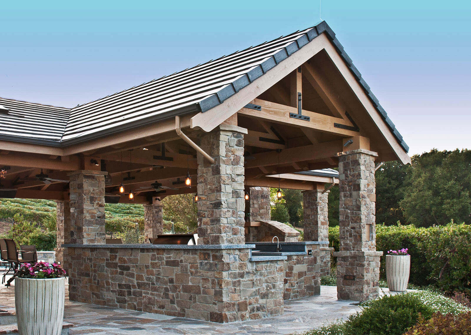 Shingled pergola with stone posts and wood rafters shelters outdoor living area, bar and fireplace with view of California hills
