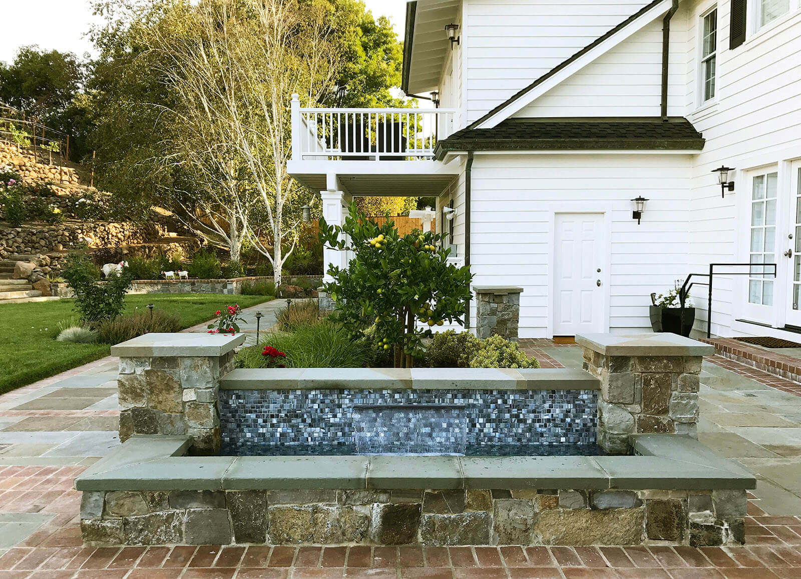Small mosaic water fountain with wrapping stone-edged pool on stone and brick outdoor patio