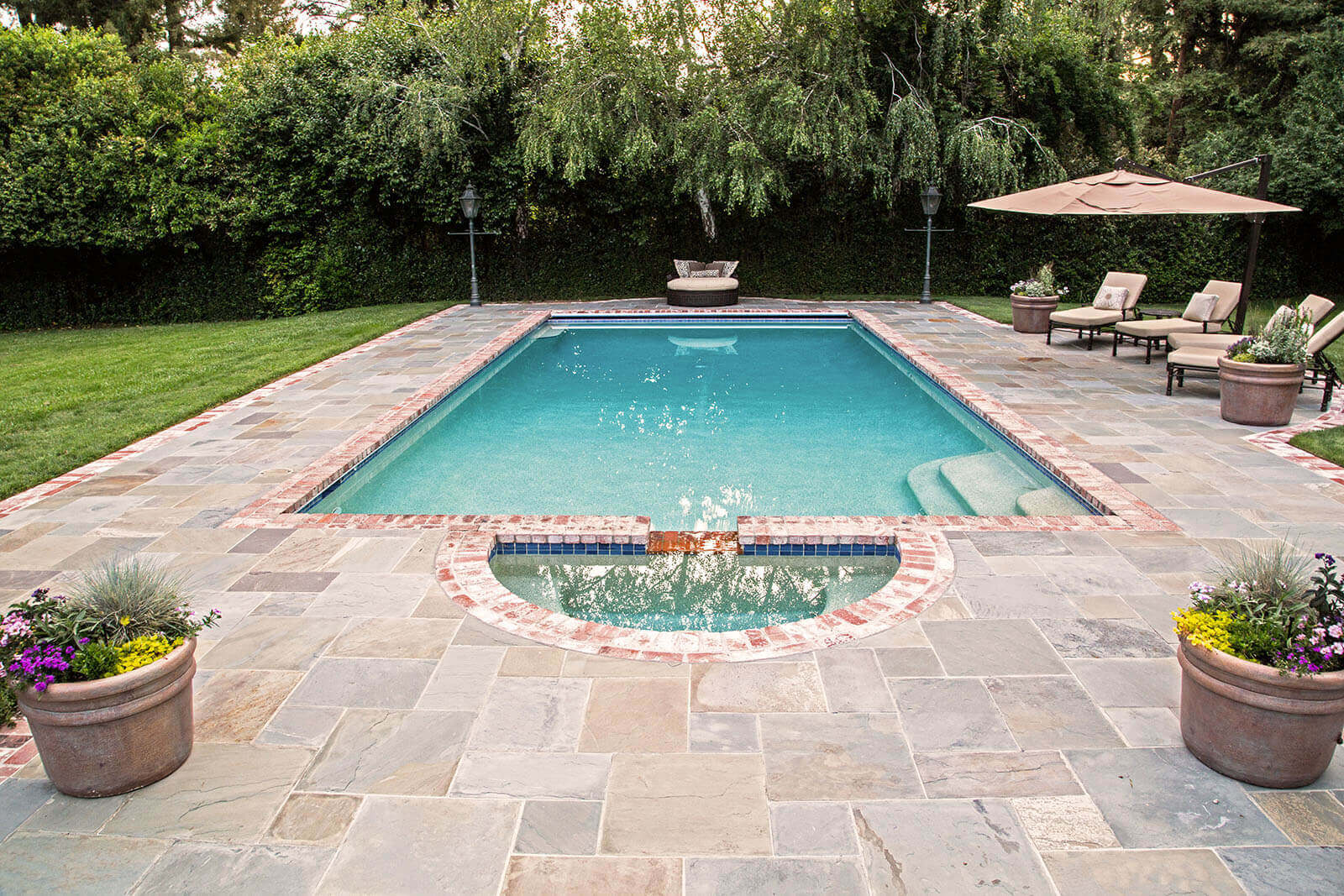 Medium-sized brick-lined aquamarine swimming pool with nearby lounging areas on slate stone tiling under tan pyramid umbrellas